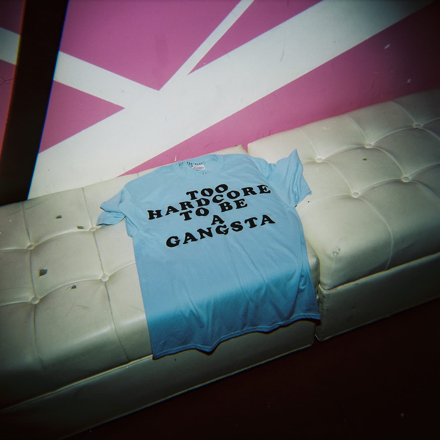 A light blue t-shirt lying on a white tufted sofa. The t-shirt has black text that reads "TOO HARDCORE TO BE A GANGSTA." The background wall is pink with white geometric lines.