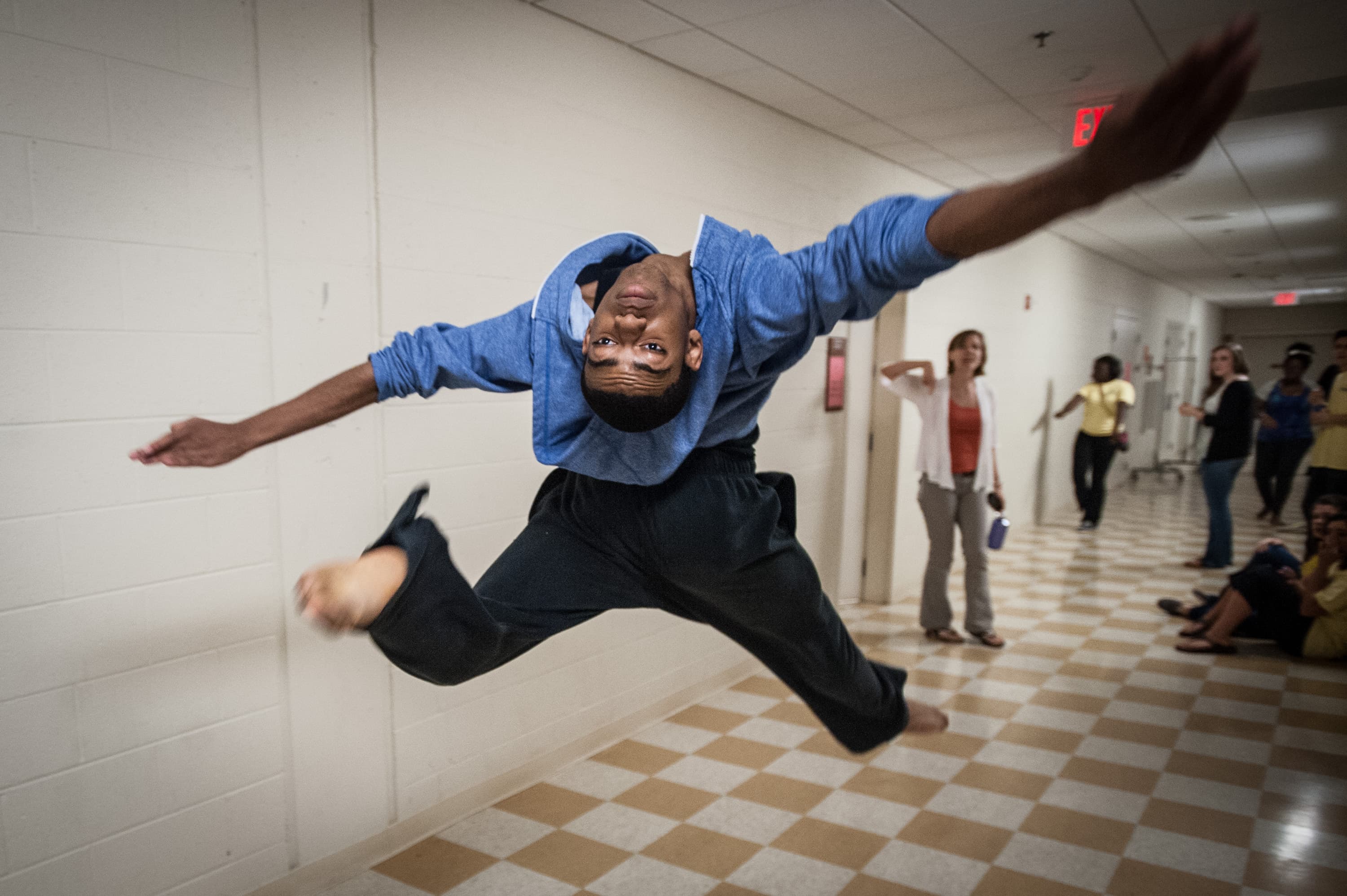 A person wearing a blue shirt and black pants is performing a high jump with a twist in a hallway. Several people in the background are observing the action.