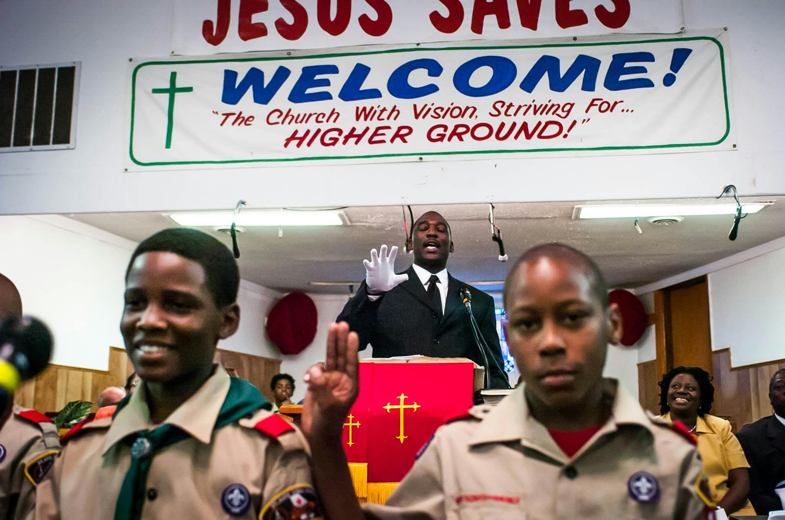 A man in a black suit wearing white glove preaching in a small church, with two boy scouts in the foreground.