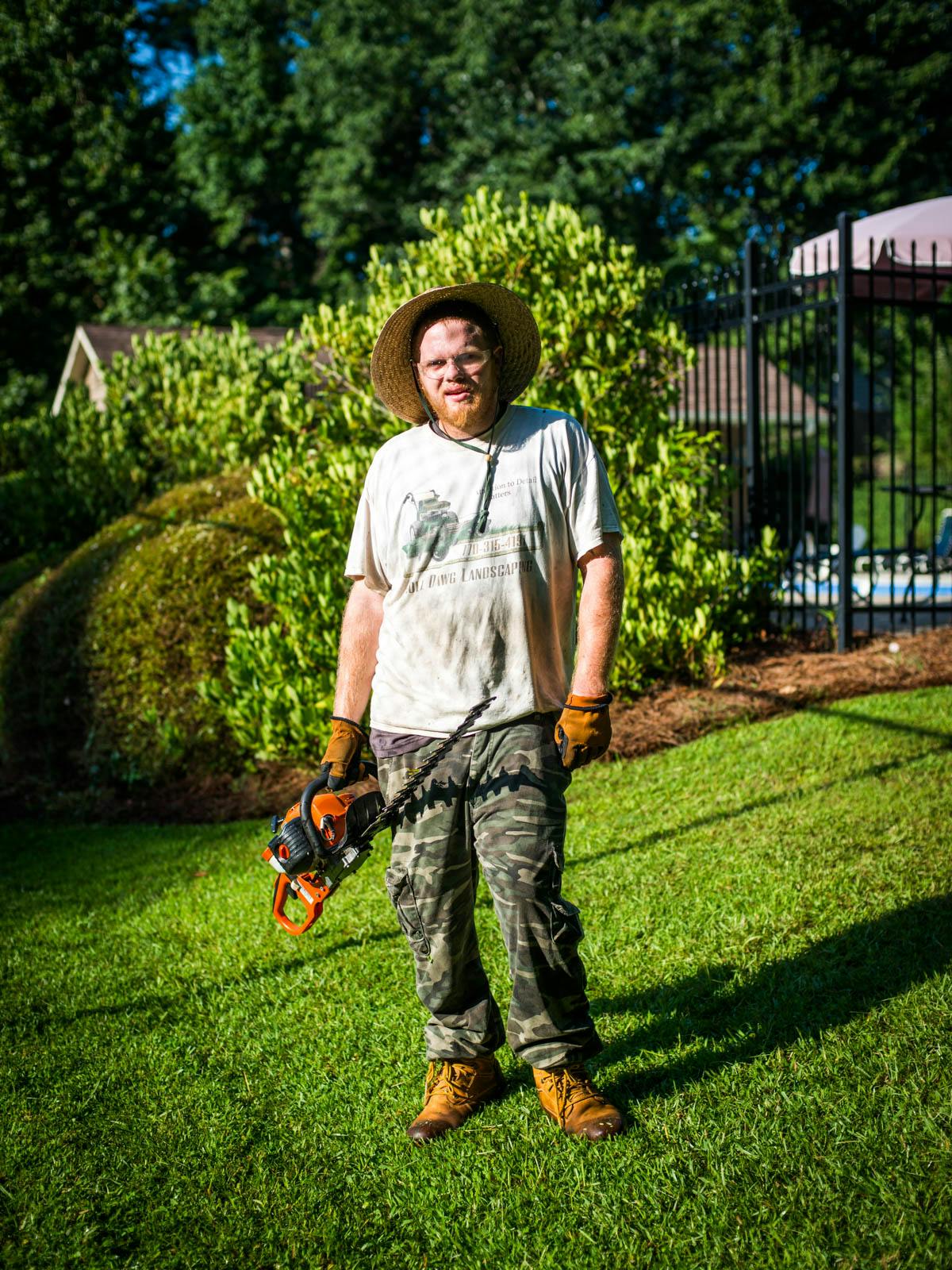 Lawn care worker holding hedge trimmer and wearing camo pants and hat, looking at the camera.