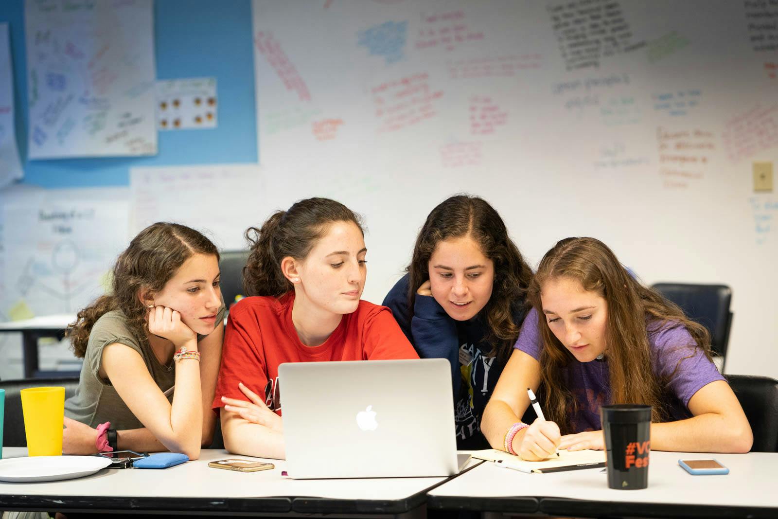 Four teenage girls huddled around a laptop working together.