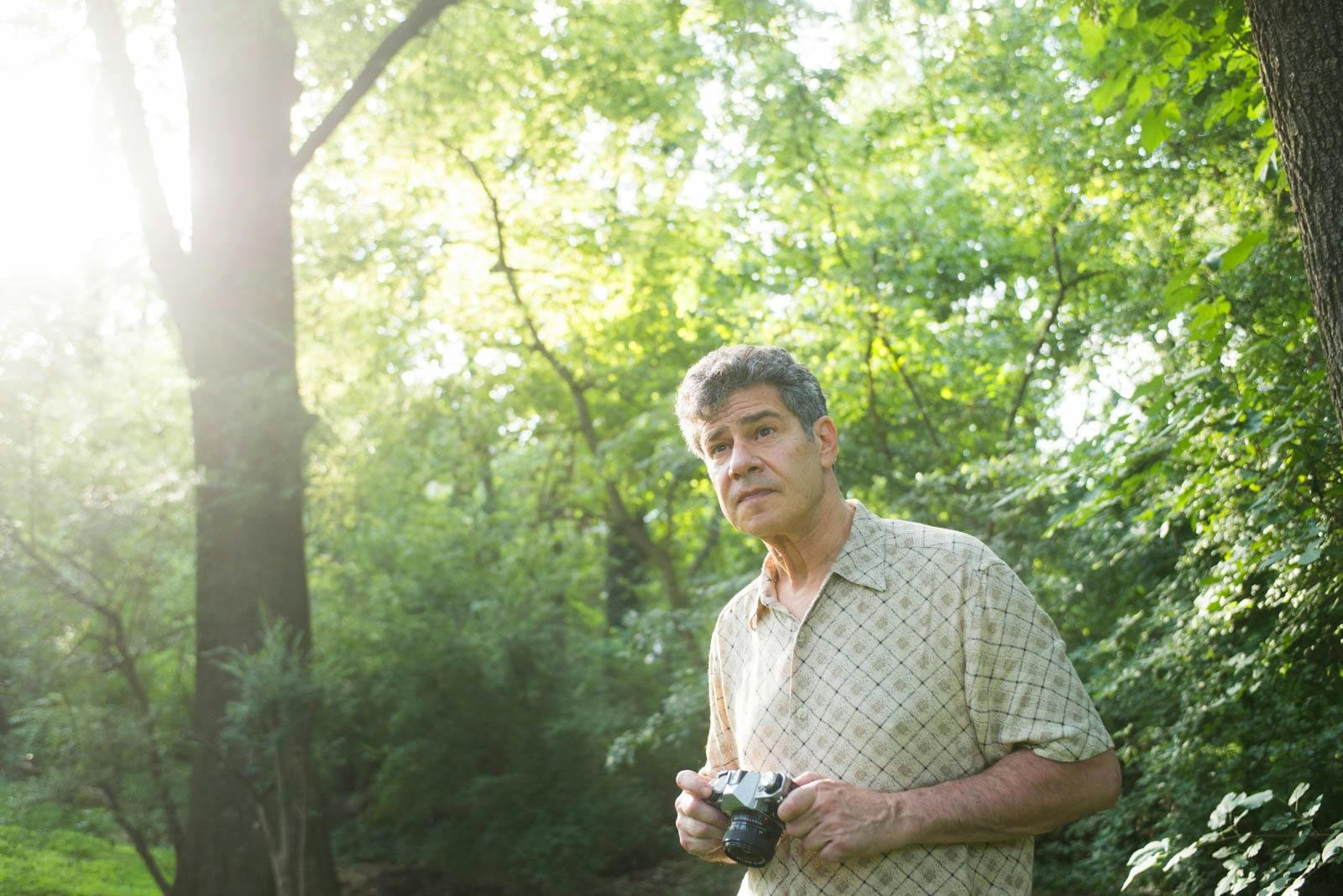 A man with grey hair in the woods holding an SLR camera.