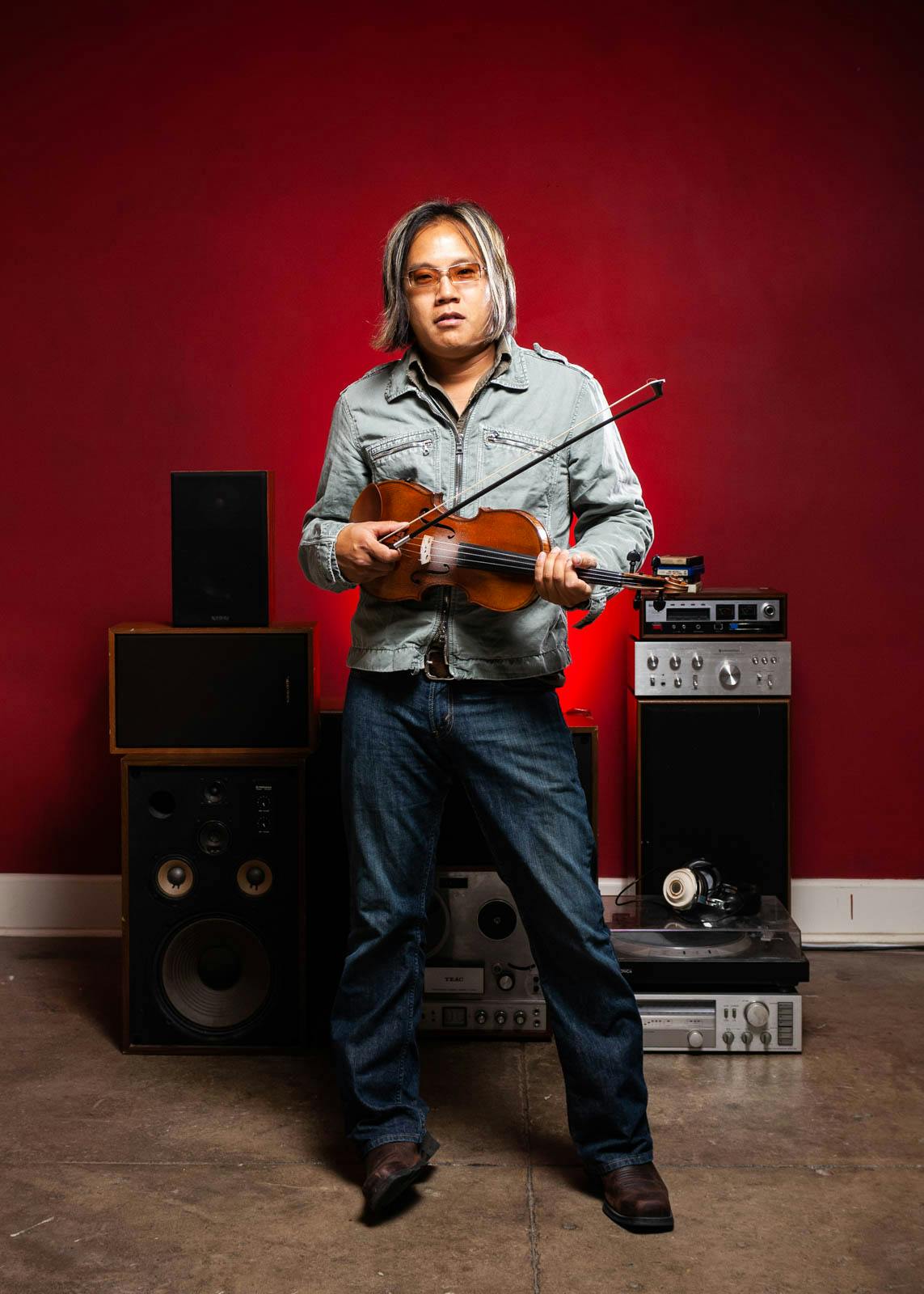 Rock and roll violinist against a red wall with amps and speakers behind him.