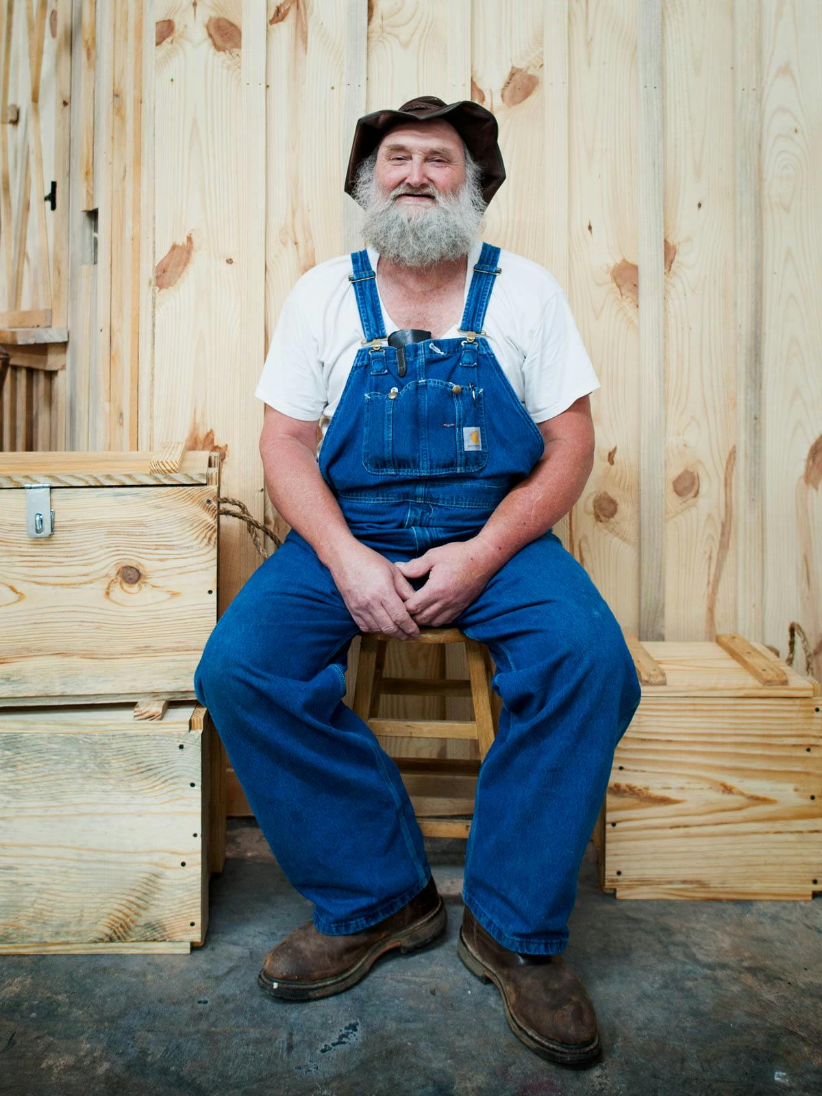 Man with shaggy beard and coveralls sitting on a stool in a distillery.